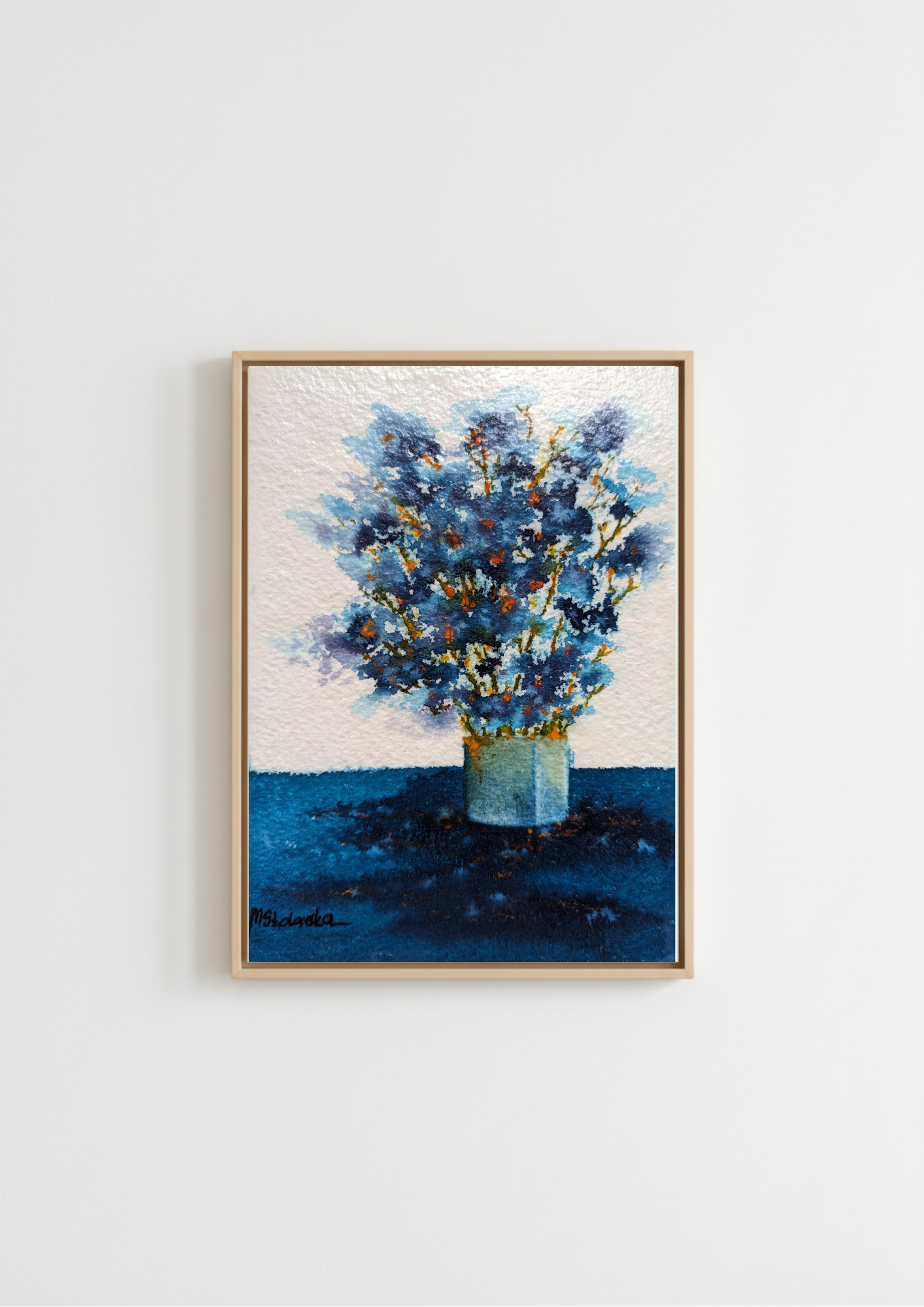 A framed painting of blue flowers in a vase, displayed on a light gray wall. The painting features textured blue and white brushstrokes, with a light wood frame.