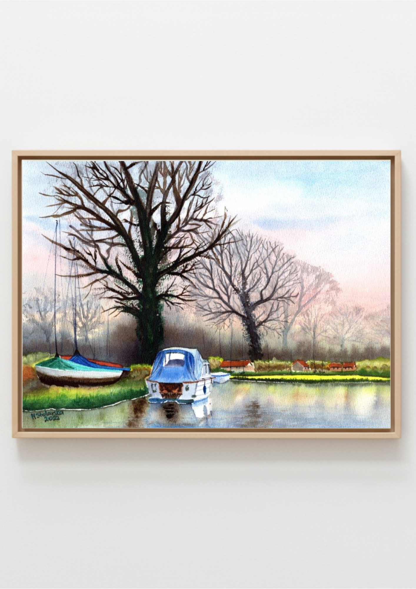 A framed watercolor landscape painting of a quiet canal scene with leafless trees, a blue boat in the foreground, and another boat on the left, shown against a light gray wall.