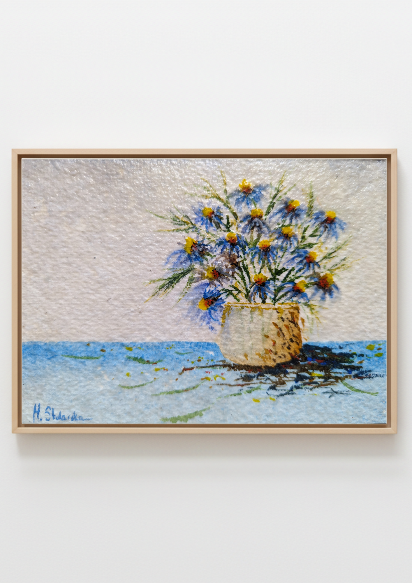 A framed painting depicting a bouquet of blue and yellow flowers in a textured pot against a light sky and blue water background, mounted on a plain white wall.