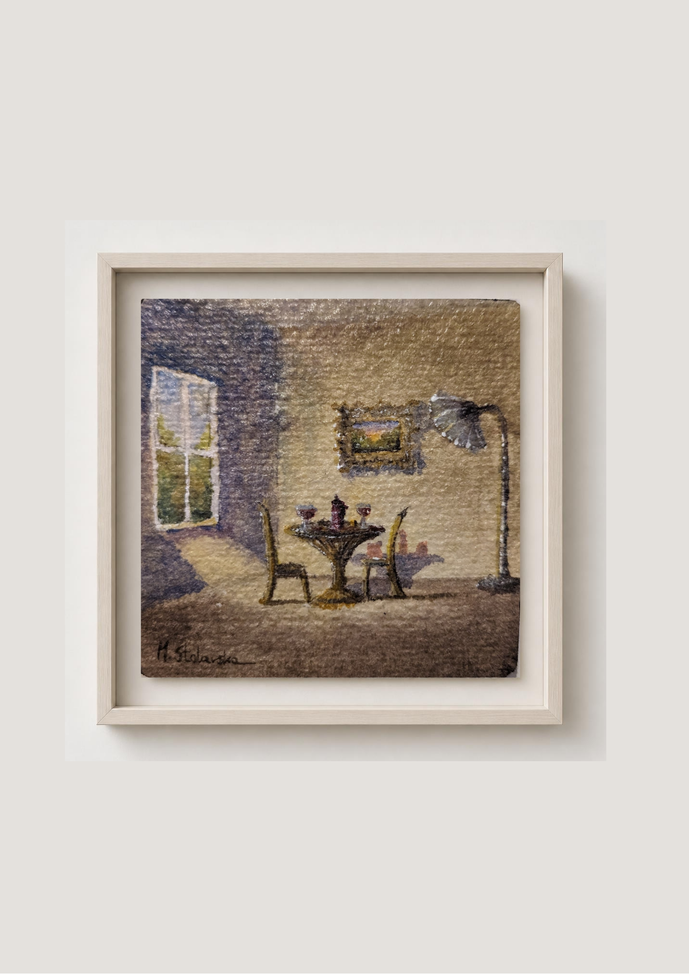A framed artwork depicting a cozy interior dining scene with a table, chairs, and a window, mounted on a light beige wall.