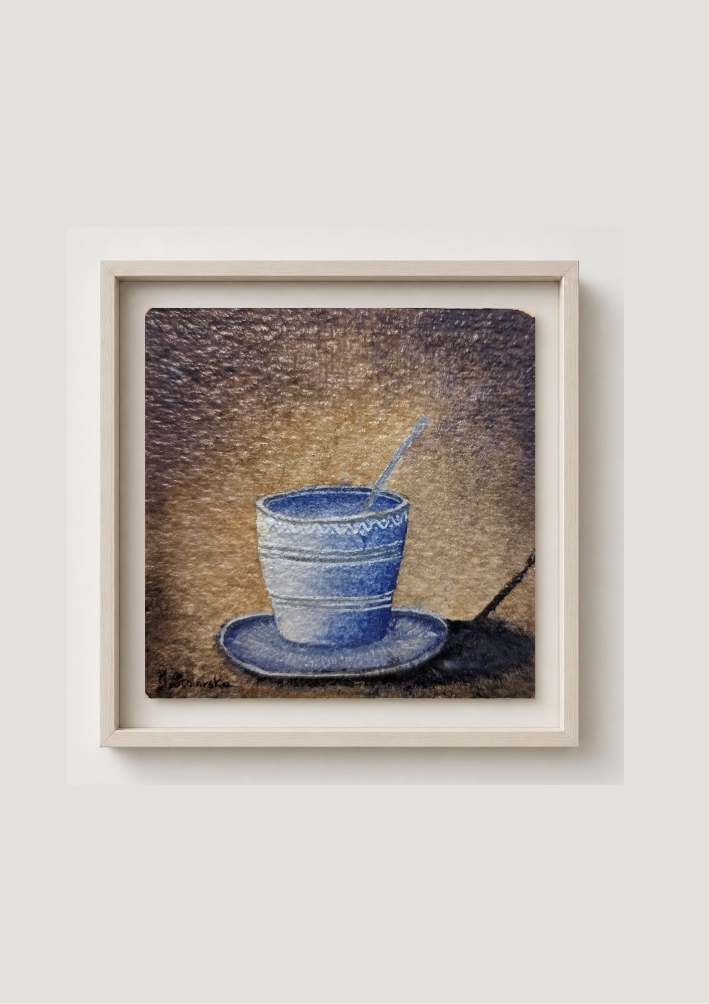 A framed art print depicting a blue cup and saucer with a spoon on a textured brown background, set in a light beige frame against a neutral light gray wall.