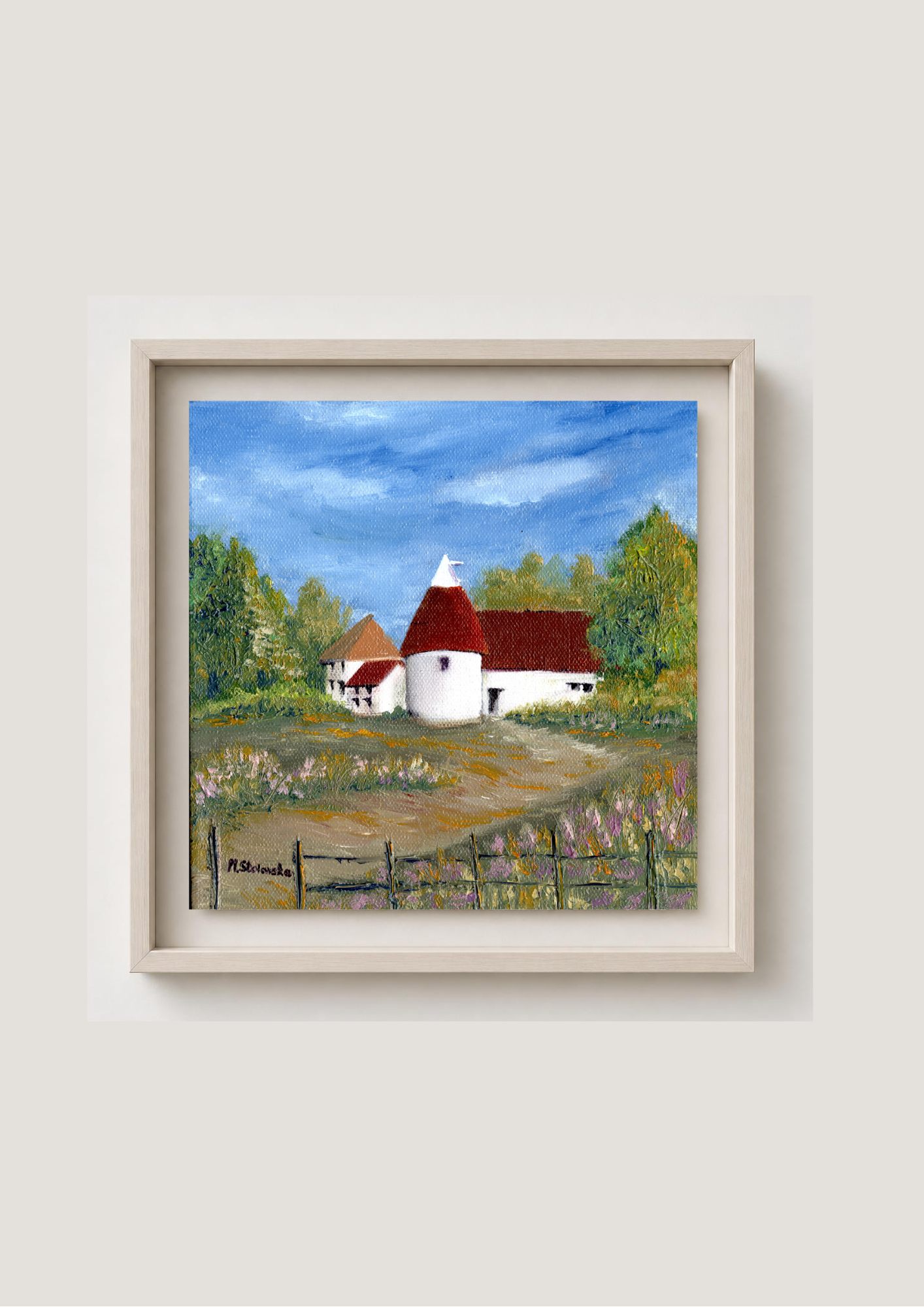 A framed landscape painting of a rural scene with white buildings and red roofs under a blue sky, displayed against a light beige wall.