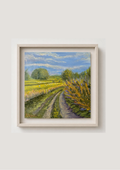 A framed landscape painting of a dirt road winding through a green field under a blue sky, with fields of sheep grazing in the background, presented in a light wood frame against a neutral beige wall.