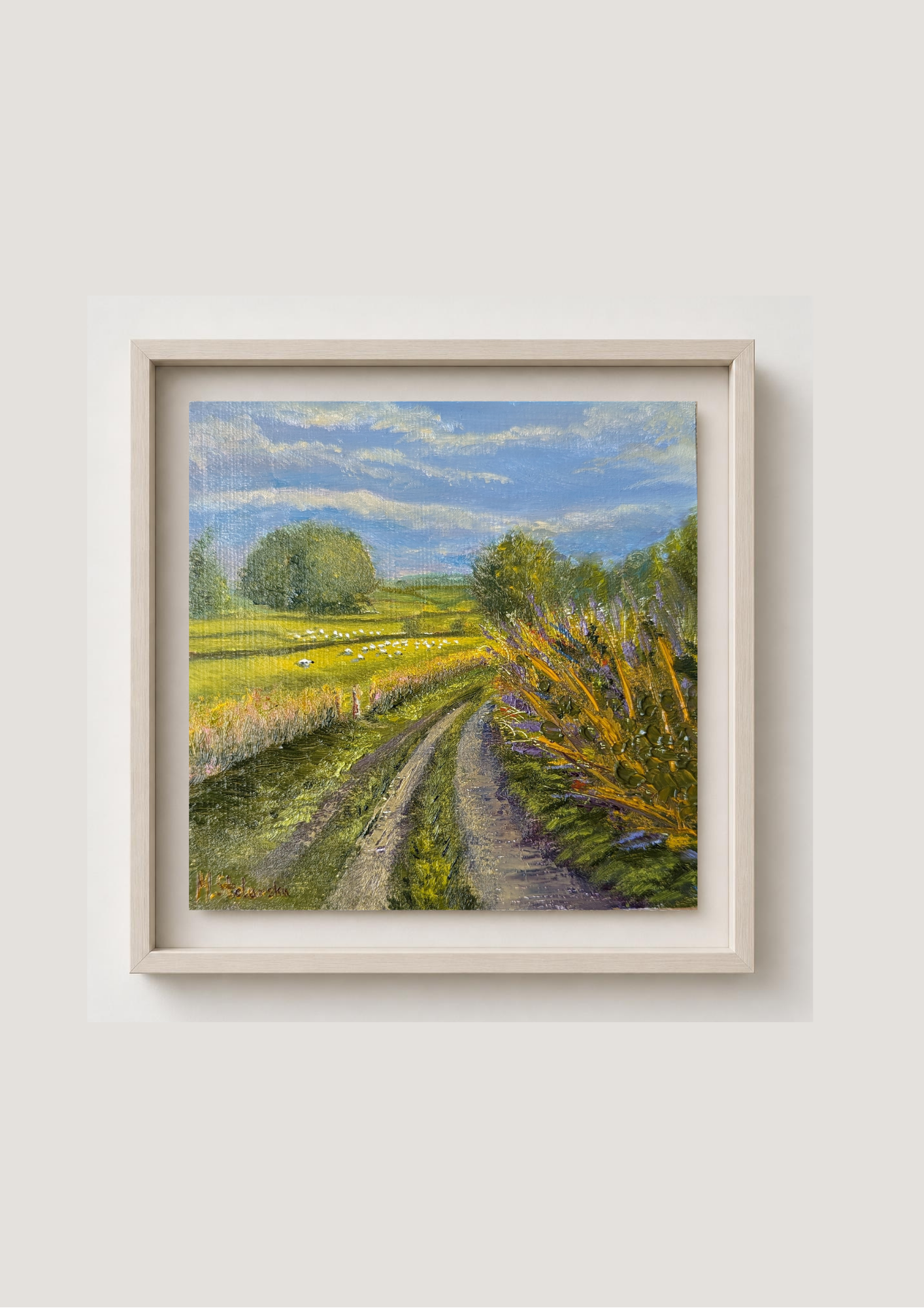 A framed landscape painting of a dirt road winding through a green field under a blue sky, with fields of sheep grazing in the background, presented in a light wood frame against a neutral beige wall.