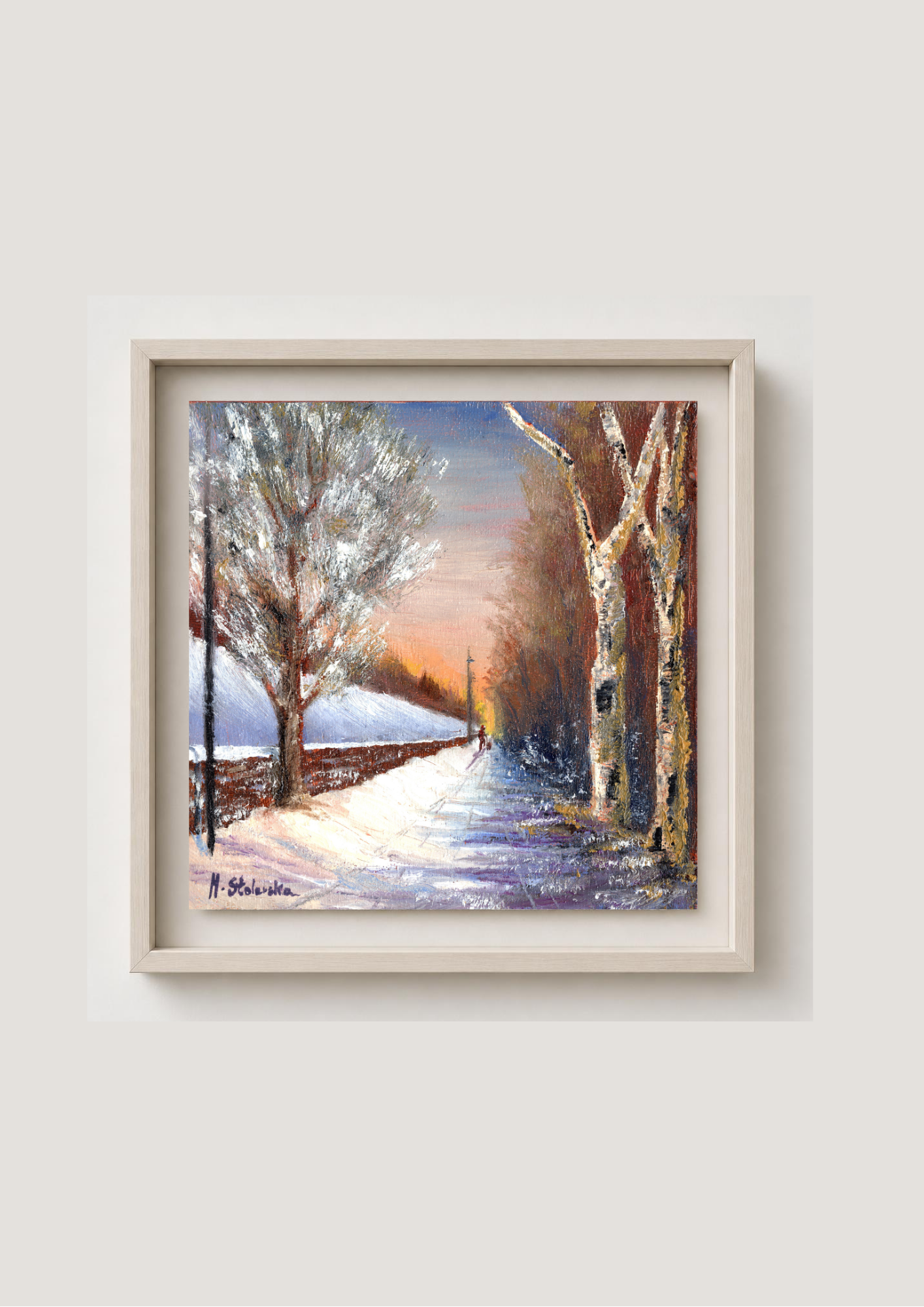 A framed winter landscape painting featuring a snow-covered path lined with leafless trees under a warm orange and blue sky, displayed on a light beige wall.